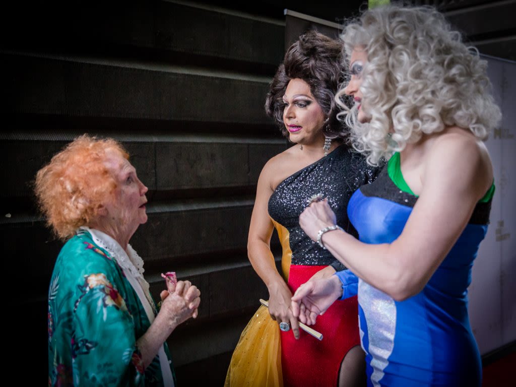 Two drag queens conversing with an older woman at the Coming Back Out Ball